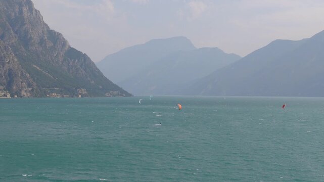 Beautiful aerial drone footage of kitesurfers riding in the middle of Lake Garda, surrounded by stunning mountains and scenic beaches. Vibrant summer atmosphere with clear blue water and dynamic motio