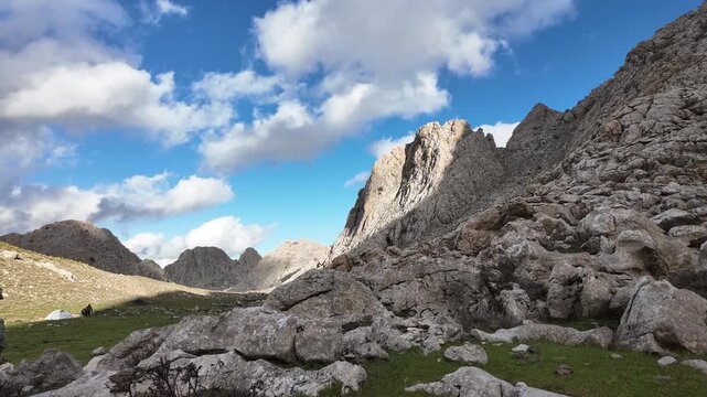 Time-lapse a wide-angle view of a rugged, rocky mountain landscape with a small camping tent visible in a green valley, set against a dramatic sky with white clouds in Tikjda, Algeria