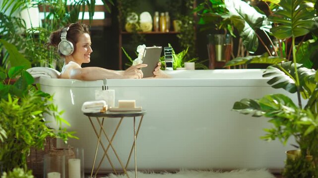 A woman smiles unwinding in a bubble bath, using a tablet & headphones in a serene, plant-filled bathroom. Luxurious setting with candles creates a peaceful, modern self-care oasis.