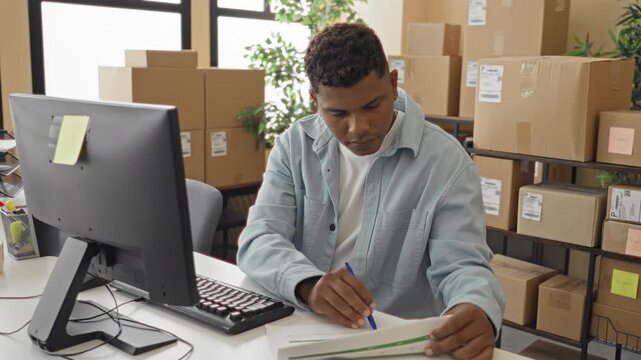 Man signing parcel form with pen and computer at desk in building surrounded by stacked cardboard boxes and shipping labels; efficiency focus.