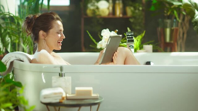 A smiling middle-aged woman enjoys a bright, relaxing bath in a plant-filled bathroom, comfortably engaging with her tablet during a peaceful self-care moment.