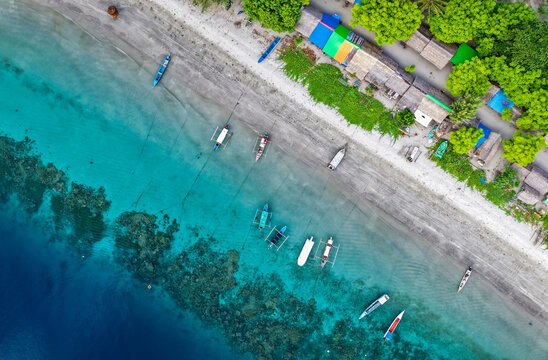 Aerial top-down view of a tropical beach with turquoise water, coral reefs, and colorful huts nestled under lush green trees. Location in Atauro Beach, Timor Leste