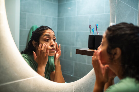 Young woman applying eye cream in home bathroom mirror
