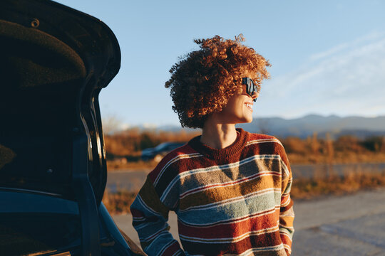 Woman lifestyle outdoors beside car with mountains at sunset curly hair profile portrait on outdoor road autumn scenery warm golden light calm mood peaceful travel moment in nature