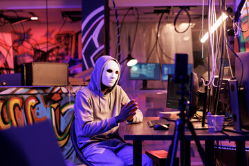 Fototapeta premium Criminal in anonymous mask recording online fraud video on smartphone. Hacker with hidden identity live streaming threat on mobile phone and asking victim for information on dark web