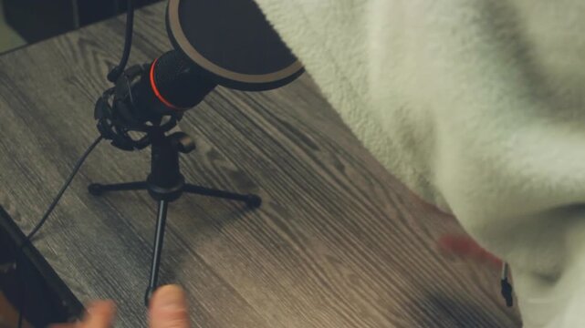 Anonymous Podcast Host In Hood Speaking Into Microphone On Wooden Desk. Close Up Overhead View Of Person Gesturing With Hands While Recording Audio Content. Professional Home Studio