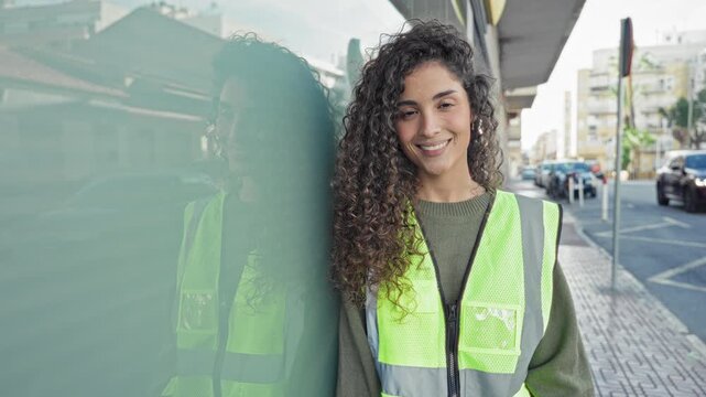 Woman in reflective vest smiles and leans against a glass wall on a busy street, construction worker; confidence safety.