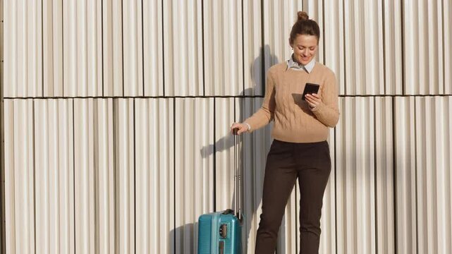 A smiling woman with her hair in a bun stands by her blue suitcase, checking her mobile phone in front of a modern white wall, ready for her next journey.