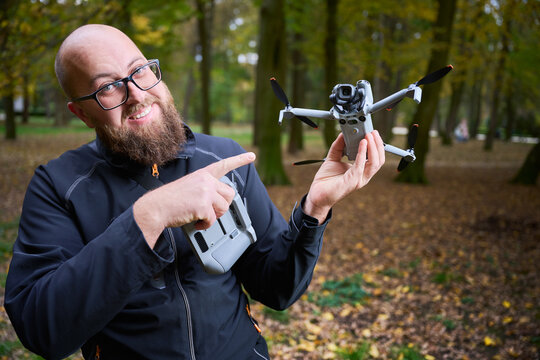 Man points at drone and shows it to camera. Smiling bearded man holding quadcopter and controller in a park, presenting equipment for aerial photography, concept of modern technology and hobby