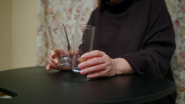 Close up of a person holding and striking two empty glasses on a black table. The woman, wearing a brown sweater, seems to be waiting or anxious, subtly moving the glasses back and forth