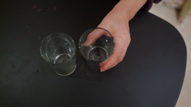 A high angle shot of a person's hand striking an empty, wet glass tumbler on a dark table. Another empty glass sits nearby. The action could suggest nervousness, contemplation, or cleaning