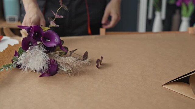 Man florist leans on table smiling with arms crossed, bouquet of purple calla lilies on brown wrapping paper in studio; friendly craft.