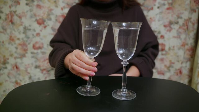 A woman sits at a dark table hitting, striking, clinking glasses, she swirls water in a vintage glass. Another full glass stands beside it, suggesting a moment of waiting, anxiety, or deep thought