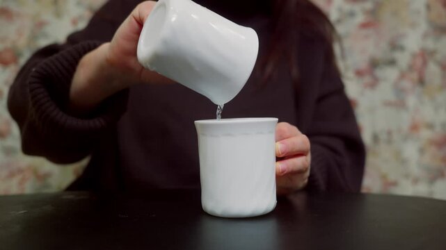 A woman sits at a black table and pours clear water back and forth between two identical white ceramic mugs. The focus is on her hands and the mugs against a blurred floral background