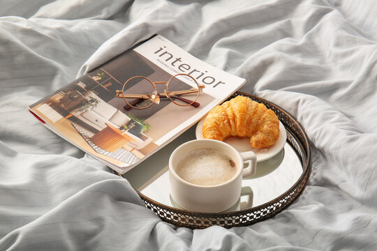 Cup of coffee with croissant, magazine and eyeglasses on light bed sheets