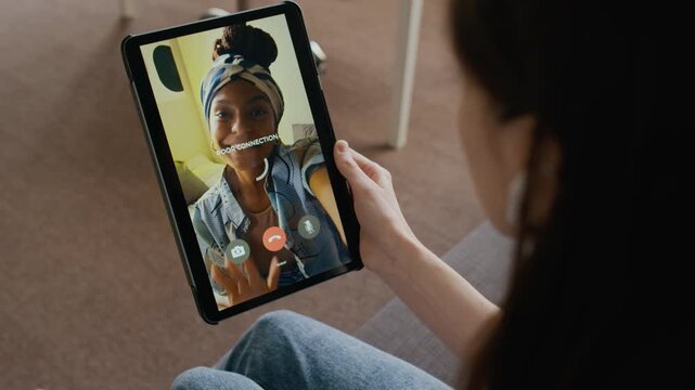 Over-shoulder shot of Caucasian woman chatting to excited African American girlfriend on video call on wireless tablet computer, experiencing internet connection problems, image freezing on screen