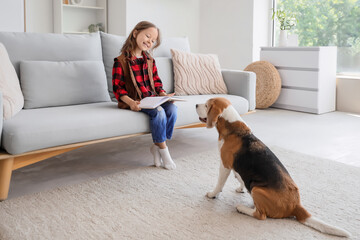 Naklejka premium Cute little girl with Beagle dog reading book on sofa in living room