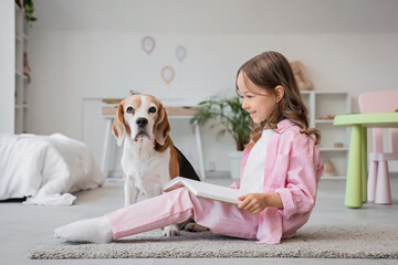 Naklejka premium Cute little girl with Beagle dog reading book on floor in bedroom