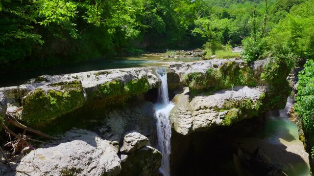 A scenic view of a powerful waterfall cascading down mossy rocks into a narrow canyon. A tranquil river flows above, surrounded by a lush green forest on a bright, sunny day. The camera tilts down
