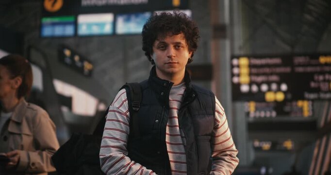 Young Traveler With Curly Hair Stands in Center of Modern Airport Departure Hall. Wearing Striped Shirt, Blue Vest, Carrying Black Backpack, Man Has Calm, Ready Expression. Modern Traveler. Portrait.