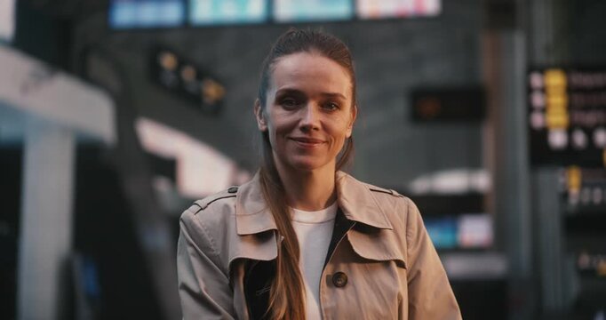 Young Woman in Beige Trench Stands Still and Smiles in Middle Busy Airport Departure Hall. While Other Passengers Walk Past in Motion Blur, Lady Remains Calm and Joyful, Looking Forward Her Journey.