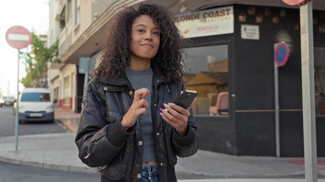 Teenage african american woman with curly hair holding smartphone, tapping screen and small fist pump on street; joy victory.