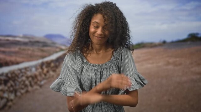 Teenage woman with curly hair making a crossed hands x gesture on a rural street by a stone wall, hands front and visible; rejection doubt.