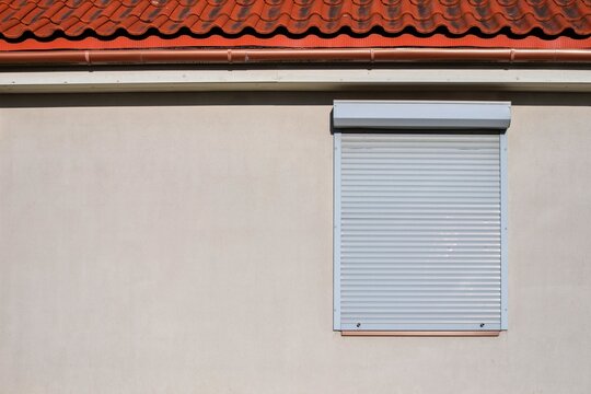 Closed white roller shutter on a house window for security and sun protection. Minimalist beige wall background with orange tile roof. Protection from burglary and heat. Copy space.