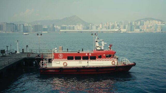 Static wide shot of Hong Kong Fireboat 8 docked at harbour pier, red and white vessel with Chinese fire department markings, Kowloon skyline and Lion Rock mountain visible in background.