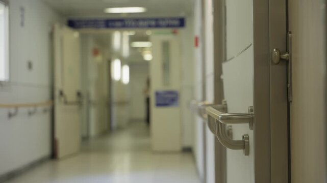Hospital corridor with person walking in background. White walls in quiet and peaceful moment inside indoor hallway in public hospital clinic. Establishing shot with focus on doctor's office door