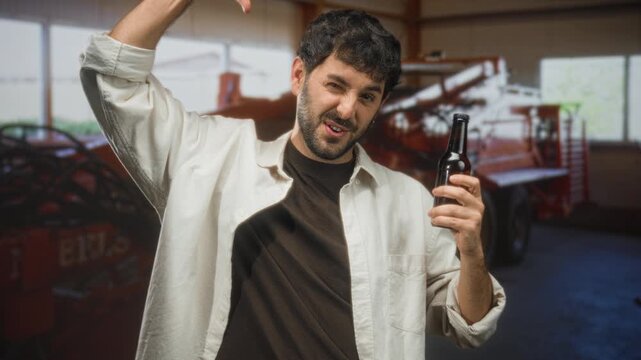 Man holding a beer bottle in right hand and raising left arm while smiling inside a workshop building with machinery and windows visible; casual cheer.