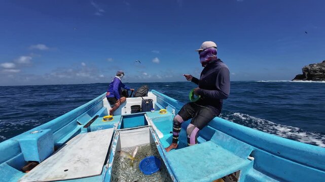 Wide shot of two local fishermen navigating choppy waters with a compartment full of fish, featuring a rocky island and seagulls in the background.