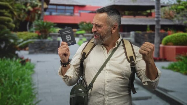 Man holding passport and fist pumping in front of a modern building with backpack and canteen visible, smiling broadly; travel excitement.