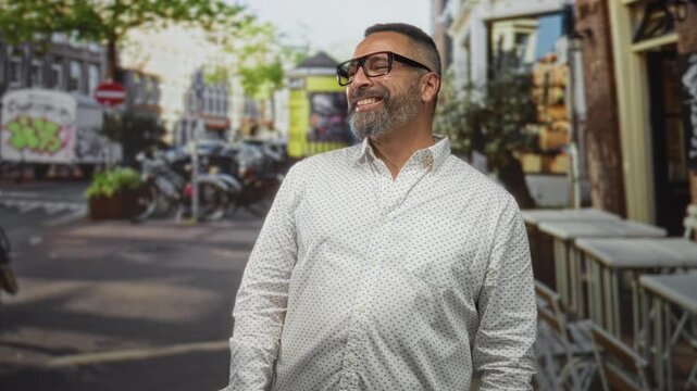 Man with beard smiling and looking to the right on a city street cafe, wearing glasses and a patterned shirt; joy confidence.