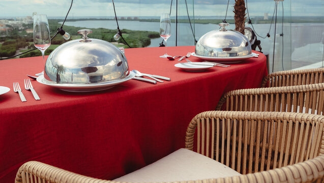 The image zooms in on an elegantly arranged dining table featuring shiny cloches and fine tableware, set against a tranquil table with nature as its peaceful backdrop.