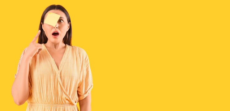Shocked young woman pointing at sticky note on her forehead against yellow background with space for text