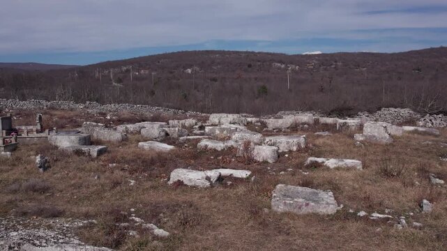 Wide view of medieval stecci tombstnes scattered across a rural landscape in Herzegovina, with hills and sky in the background.