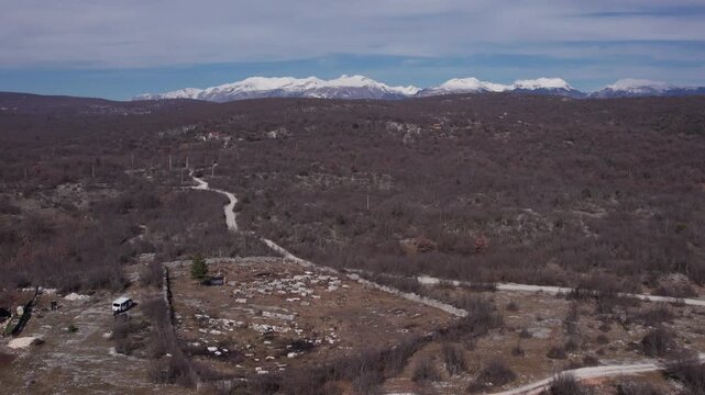 Drone shot of medieval stecci necropolis in Herzegovina with rural landscape and snow-capped mountains in the distance.