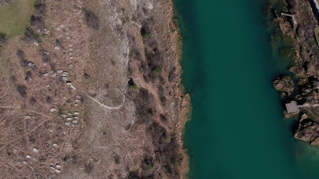 Top-down drone view of historic necropolis with scattered stone tombs near a turquoise river, set in a dry Herzegovina landscape.