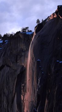 Vertical Screen: Yosemite National Park Firefall phenomenon with waterfall glowing orange and red against dark granite cliff face, snow patches dotting rugged rock texture