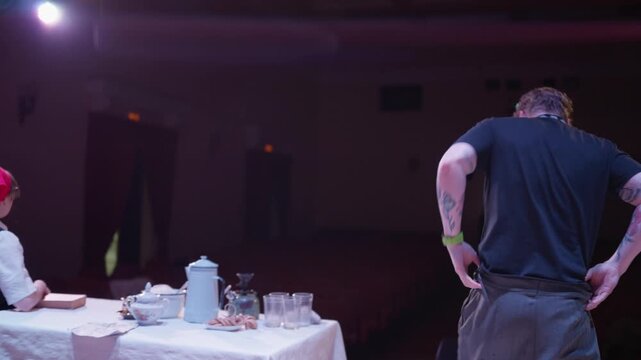 Intimate stage performance with two actors at a table in an empty theater, featuring dramatic lighting, quiet dialogue, and a minimal set design focus