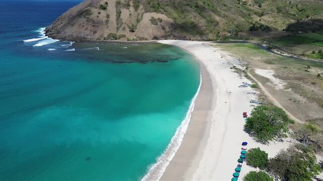 Aerial view of the pristine Mawun Beach in Lombok, Indonesia, featuring turquoise crystal clear waters, white sandy coastline, and lush tropical hills under a bright sunny sky.
