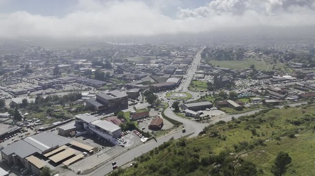 Drone flies to left from hill with parliament overlooking a roundabout on the main road, below light cloud cover on a sunny morning in Maseru, Lesotho