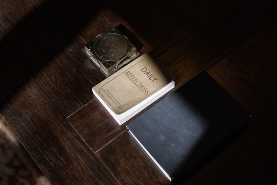 Daily Reflections book for AA members resting on a wooden table bathed in dramatic natural sunlight, casting strong shadows across the surface in a contemplative morning scene.
