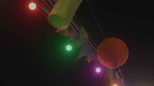 A static shot of colorful triangular bunting flags and an orange honeycomb paper lantern illuminated by glowing string lights against a dark night sky during a festive event.
