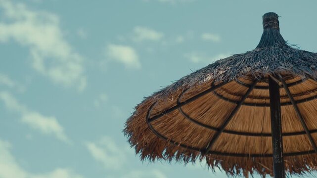 Thatched umbrella offering shade under a bright blue sky with clouds, evoking a summer vacation