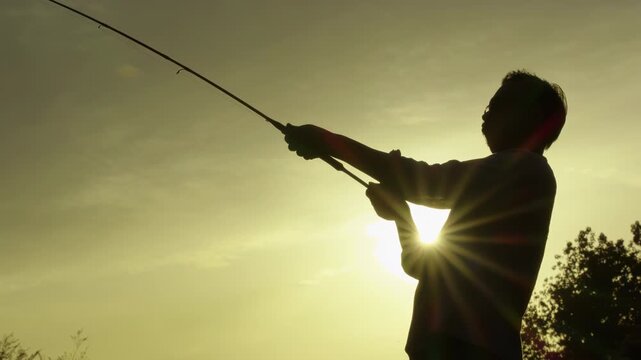Silhouette fisherman hold fishing rod with spinning reel attached, angler pulling hard after catching a big fish on the lake at, showcase peaceful outdoor leisure activity hobby recreational