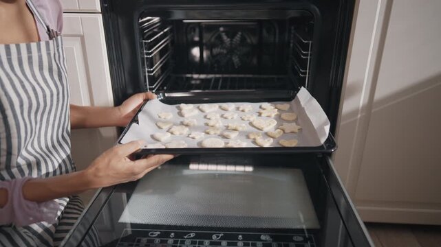 Use modern kitchen appliance, housewife holding baking sheet with raw cookies. Woman putting it into electric oven, cooking and baking tasty homemade meals for family, details shot in home kitchen
