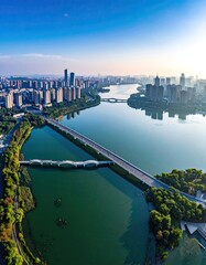 Naklejka premium An aerial view showcases a modern cityscape reflected in a tranquil river, spanned by elegant bridges under a clear blue sky.