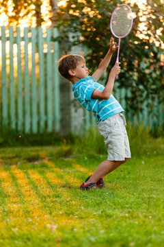 A young boy leans back dramatically as he serves the shuttlecock during an outdoor badminton game in summer. Active play, coordination, and fun vacation moments in a green field.